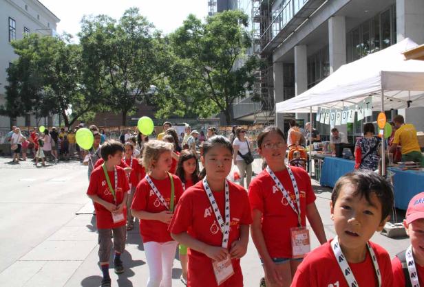 Eine Gruppe Kinder in roten T-Shirts mit Ballons und Ausweisen vor einem Infostand.