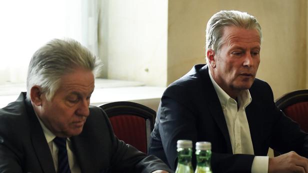 Zwei Männer in Anzügen sitzen an einem Tisch mit Wasserflaschen.