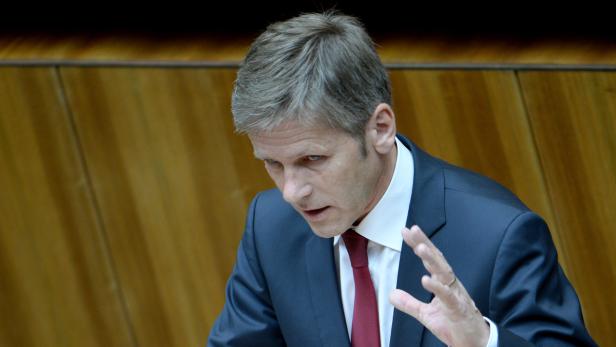 Ein Mann in Anzug und Krawatte gestikuliert vor einer hölzernen Wand.