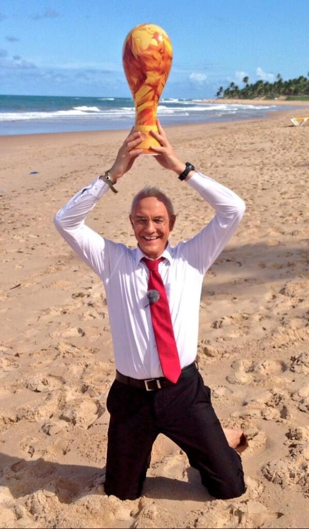 Ein Mann kniet am Strand und hält eine orangefarbene Vase über seinen Kopf.