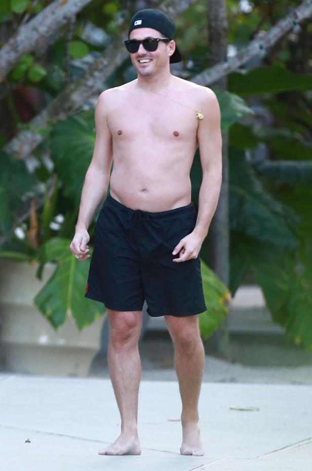 Ein Mann mit Badehose, Basecap und Sonnenbrille steht barfuß im Freien.