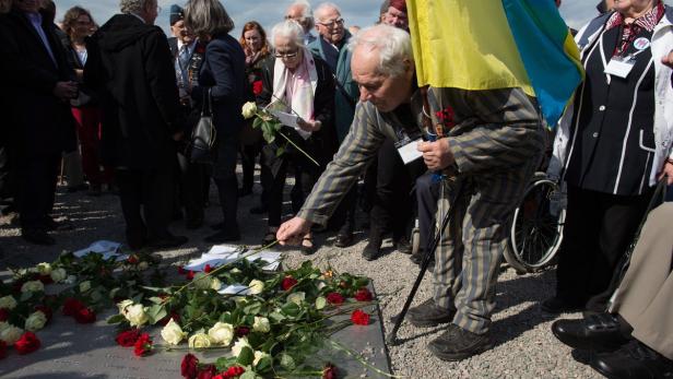 Ein Mann in gestreifter Kleidung legt eine Rose auf ein Denkmal, umgeben von anderen Menschen.