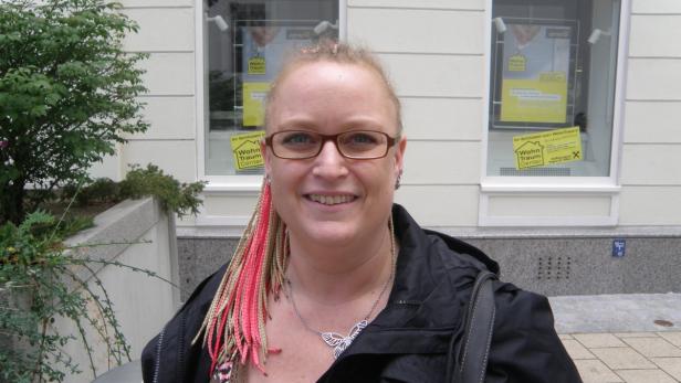 Eine Frau mit Brille und gefärbten Zöpfen steht vor einem Gebäude.