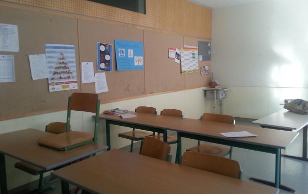 Ein Klassenzimmer mit Tischen, Stühlen und einer Pinnwand mit Postern.
