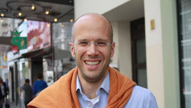 Ein lächelnder Mann mit Brille und einem orangefarbenen Pullover um die Schultern steht im Freien.