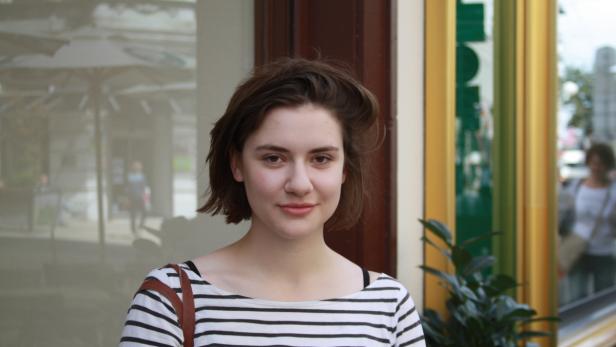 Eine junge Frau mit braunen Haaren und einem gestreiften Oberteil steht vor einem Schaufenster.