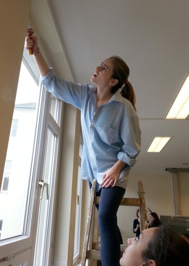 Eine Frau streicht auf einer Leiter stehend einen Fensterrahmen.