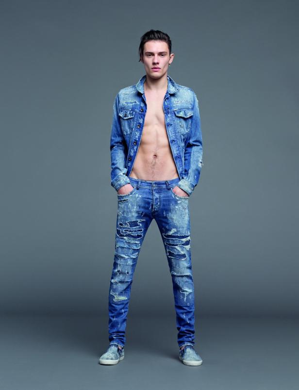 Ein Mann mit offener Jeansjacke und zerrissener Jeans steht vor einem grauen Hintergrund.