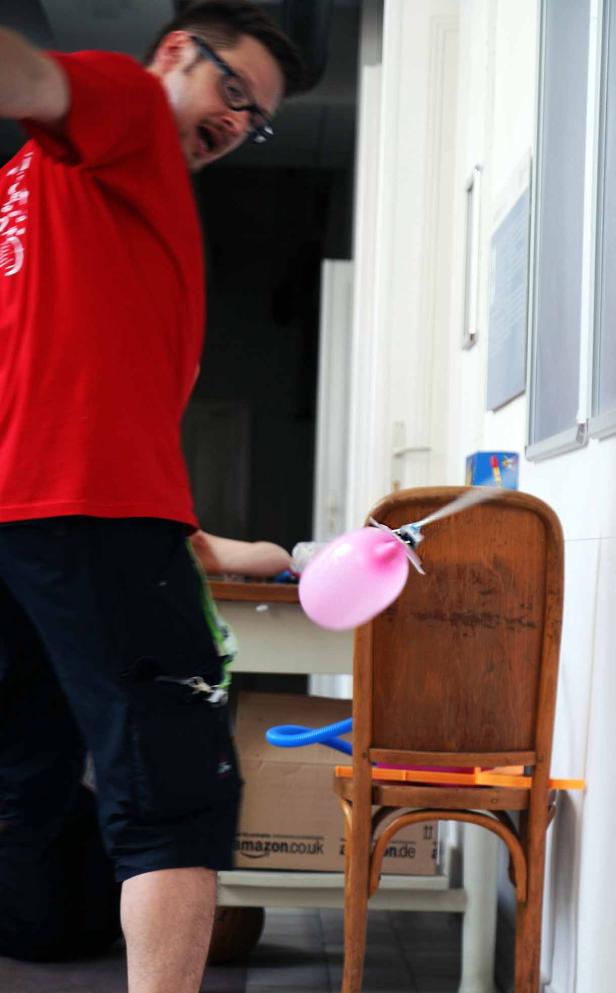 Ein Mann mit Brille versucht, einen pinkfarbenen Ballon an einem Stuhl zu befestigen.