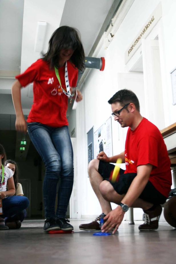 Eine junge Frau springt auf eine rote Matte, während ein Mann daneben hockt und zusieht.