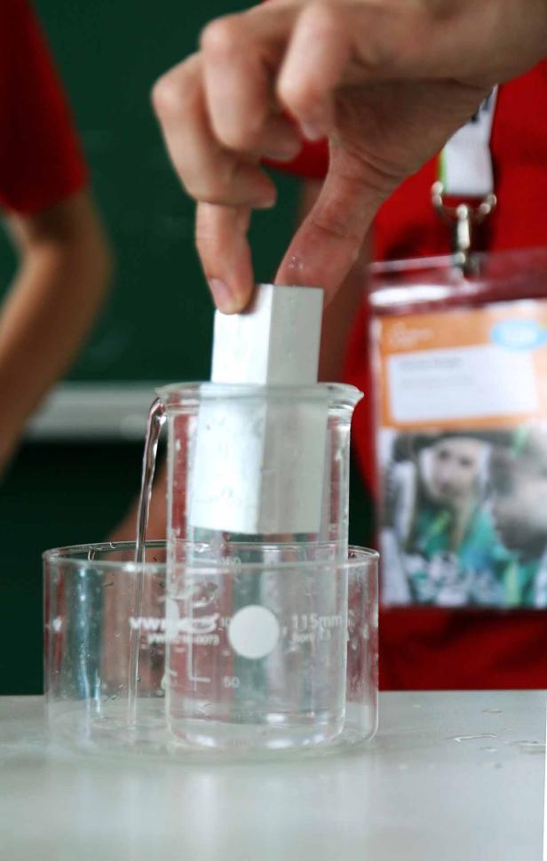 Eine Hand taucht einen weißen Würfel in ein mit Wasser gefülltes Becherglas.