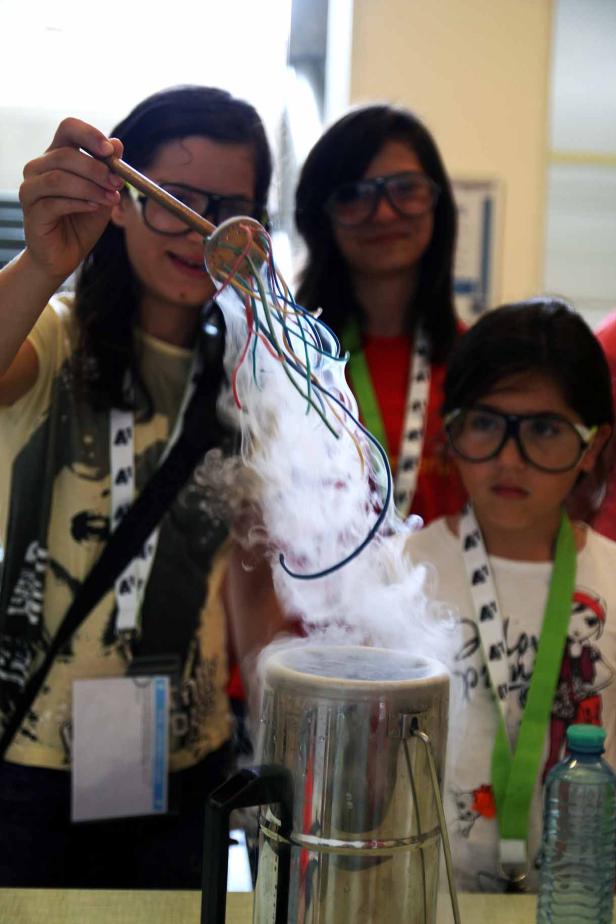 Drei Mädchen experimentieren mit Trockeneis und bunten Kabeln, wodurch Rauch entsteht.