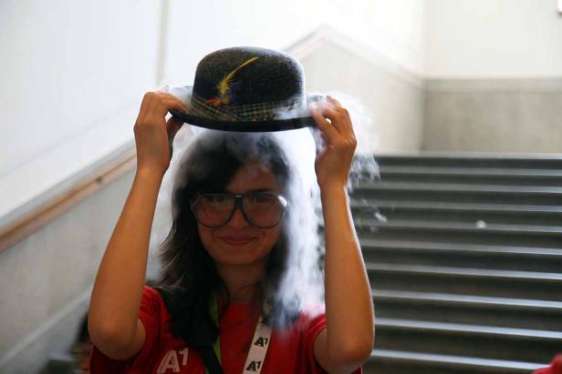 Eine Frau mit Brille hält einen Hut über ihren Kopf, aus dem Rauch aufsteigt.