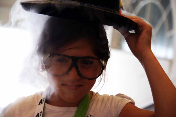Ein Mädchen mit Brille hält einen Hut über dem Kopf, aus dem Rauch aufsteigt.