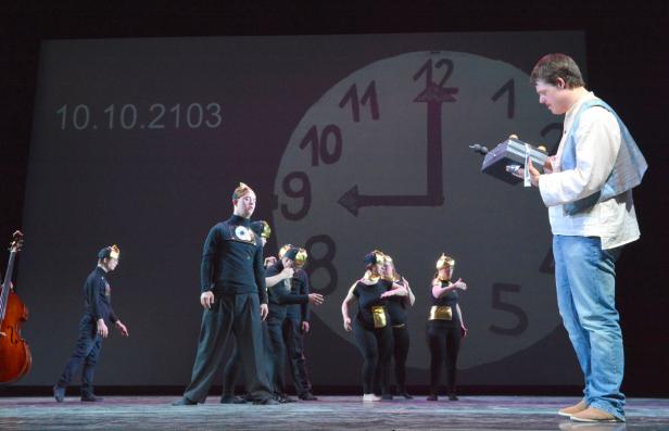 Eine Gruppe von Darstellern auf einer Bühne vor einer Uhr und dem Datum 10.10.2103.