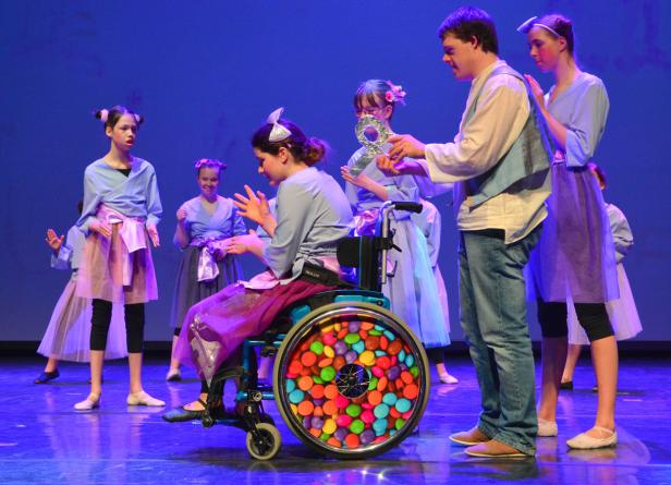 Eine junge Frau im Rollstuhl nimmt an einer Tanzaufführung mit anderen Tänzern auf einer Bühne teil.