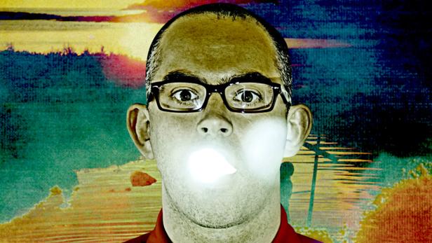 Ein Mann mit Brille bläst eine leuchtende Kaugummiblase vor einem gemalten Hintergrund.