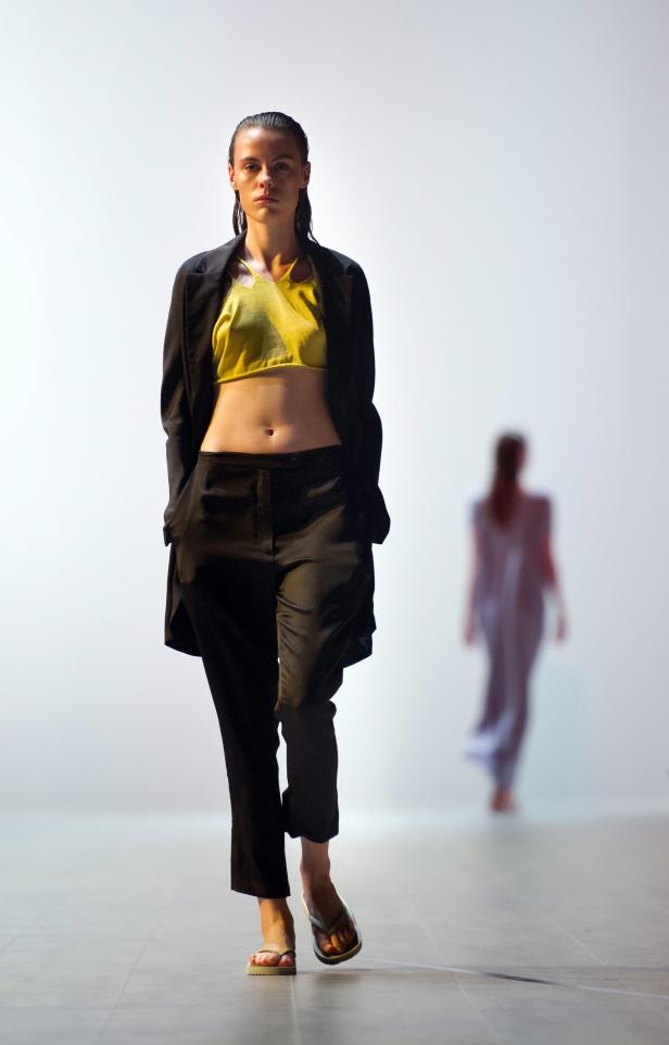 Eine Frau in einem gelben Top und einem schwarzen Blazer läuft auf einem Laufsteg.