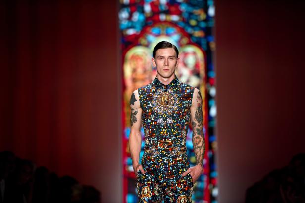 Ein tätowiertes Model mit einem farbenfrohen Outfit auf einem Laufsteg.