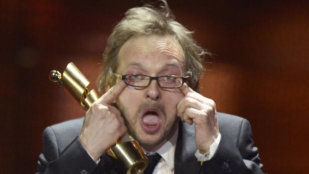 Milan Peschel spielt die Hauptrolle in "Halt auf freier Strecke". Ein Mann mit Brille verzieht sein Gesicht und hält eine goldene Trophäe.