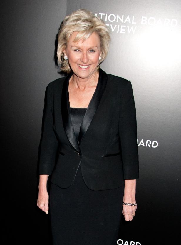 Eine Frau mit blonden Haaren lächelt in einem schwarzen Kleid und Blazer.