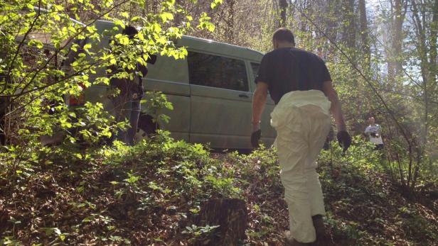 Ein Mann in Schutzanzug geht in einem Waldstück auf einen grünen Lieferwagen zu.
