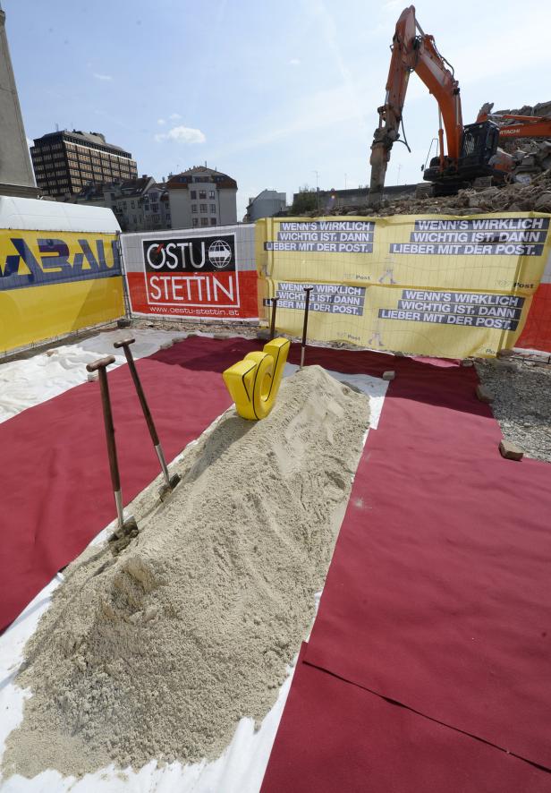 Eine Baustelle mit einem Sandhaufen, zwei Spaten und einem Bagger im Hintergrund.
