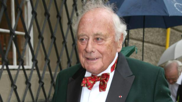 Ein älterer Mann mit Fliege und grünem Jackett vor einem Gebäude.