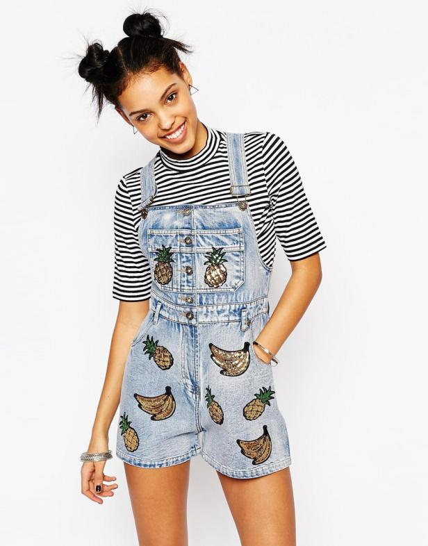 Eine lächelnde Frau trägt eine Jeans-Latzhose mit Ananas- und Bananenapplikationen.