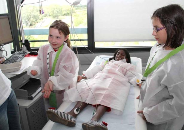 Ein Mädchen liegt im Krankenhausbett, während zwei andere Mädchen in Arztkitteln daneben stehen.