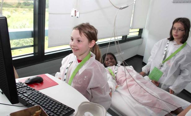 Drei Mädchen in Arztkitteln in einem Krankenzimmer, eines liegt im Bett.