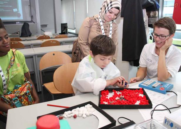 Ein Kind in einem weißen Kittel spielt mit roten und weißen Objekten, beobachtet von zwei Frauen.