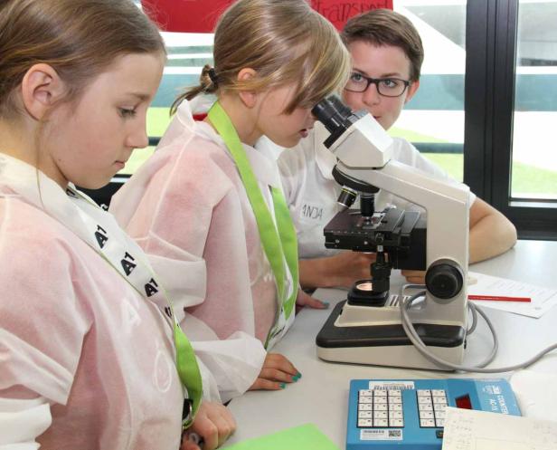 Zwei Mädchen betrachten etwas durch ein Mikroskop, während eine Frau zusieht.