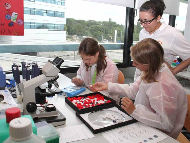 Zwei Mädchen experimentieren mit einer Lehrerin in einem Labor mit Mikroskop und bunten Kügelchen.