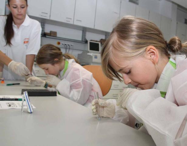 Zwei Mädchen im Labor untersuchen Reagenzgläser mit Schutzhandschuhen und Laborkitteln.