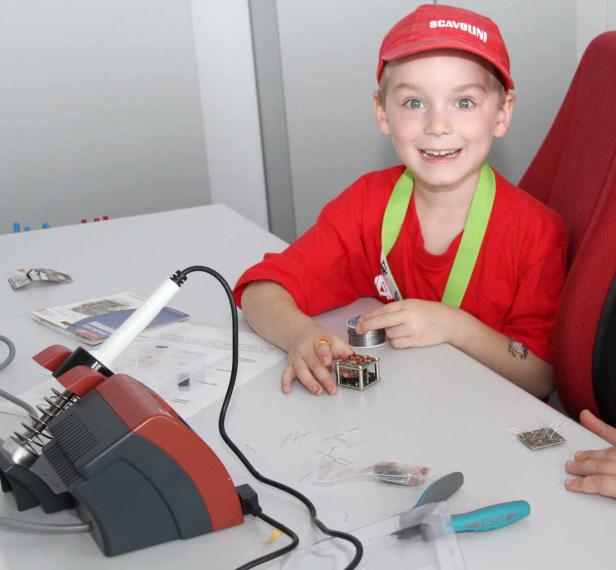 Ein lächelnder Junge sitzt an einem Tisch mit Elektronikbauteilen und einem Lötkolben.