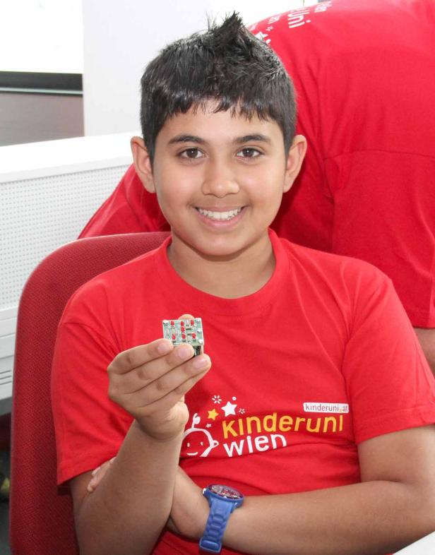 Ein lächelnder Junge mit einem T-Shirt der Kinderuni Wien hält eine Platine in der Hand.