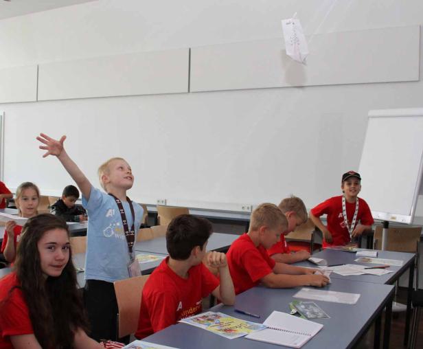 Eine Gruppe Kinder sitzt in einem Klassenzimmer, ein Junge wirft ein Papierflugzeug in die Luft.