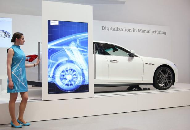 Eine Frau steht neben einem Bildschirm mit der Röntgenansicht eines Autos, daneben ein weißes Auto.