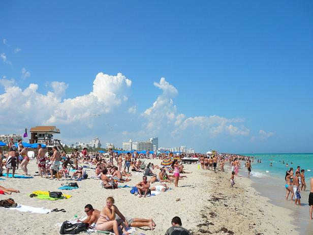 Ein belebter Strand mit vielen Menschen unter blauem Himmel und weißen Wolken.