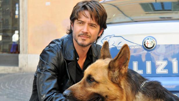 Ein Mann in Lederjacke posiert mit einem Schäferhund vor einem Polizeiauto.