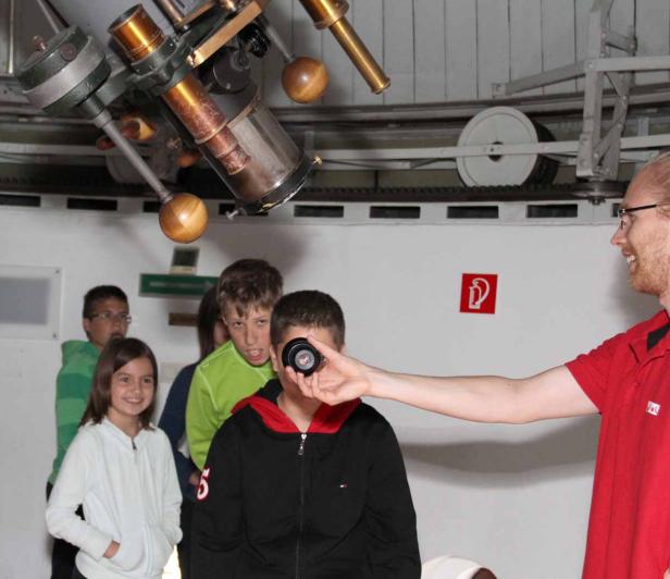 Eine Gruppe Kinder steht unter einem großen Teleskop, während ein Mann ihnen eine Linse zeigt.