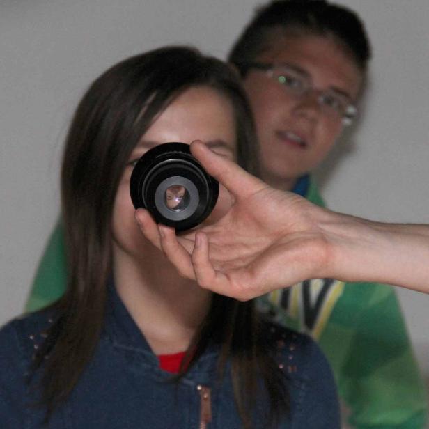 Ein Mädchen hält eine Kameralinse vor ihr Auge, während ein Junge im Hintergrund steht.