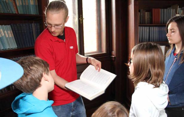 Ein Mann mit Brille zeigt Kindern ein aufgeschlagenes Buch in einer Bibliothek.