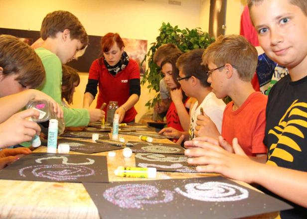 Eine Gruppe Kinder bastelt mit Glitzer und Klebstoff an Tischen.