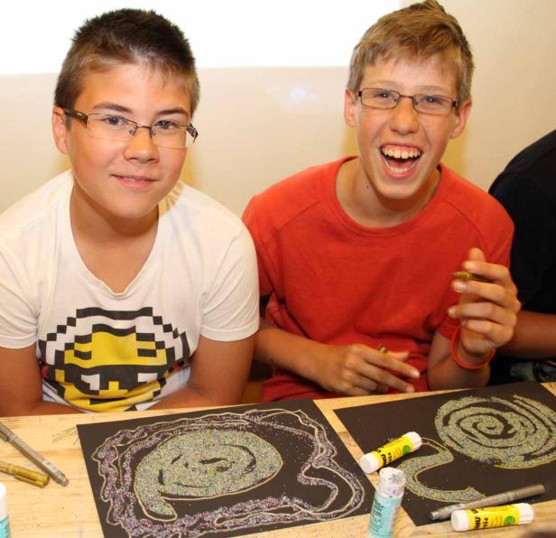 Zwei Jungen sitzen an einem Tisch und basteln mit Glitzer auf schwarzem Papier.