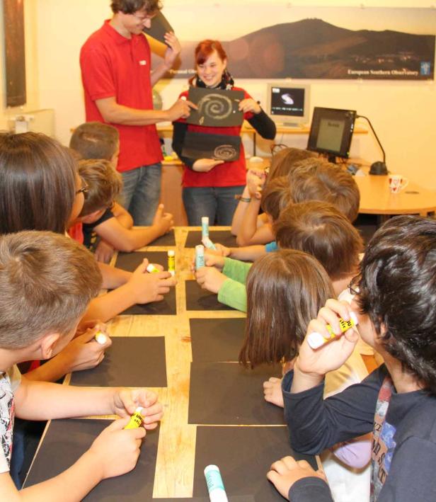 Kinder basteln mit Klebestiften auf schwarzem Papier in einem Klassenzimmer.