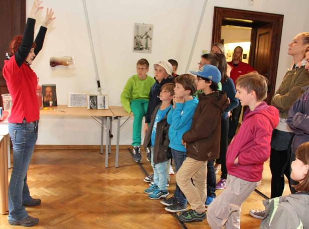 Eine Frau erklärt einer Gruppe von Kindern etwas vor einem Tisch mit Bildern.