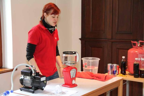 Eine Frau steht an einem Tisch mit Experimentierutensilien und einem Heliumtank.