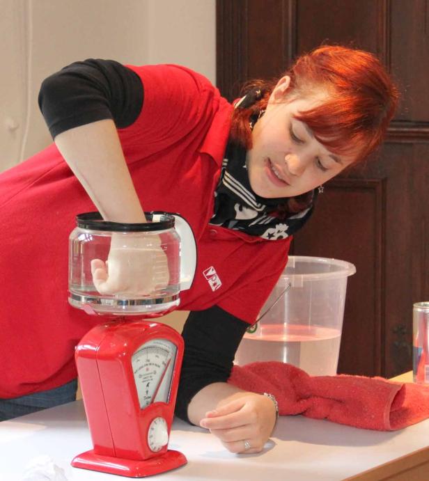 Eine Frau taucht ihre Hand in ein mit Wasser gefülltes Gefäß, das auf einer roten Waage steht.